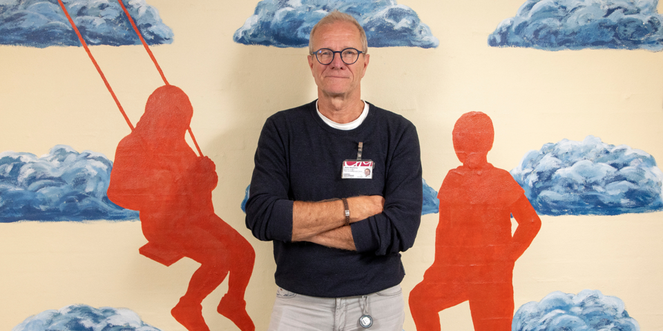 På billedet ses børne- og ungdomspsykiater Niels Bilenberg. Han står foran en væg dekoreret med malede grå skyer og orange silhuetter af mennesker  