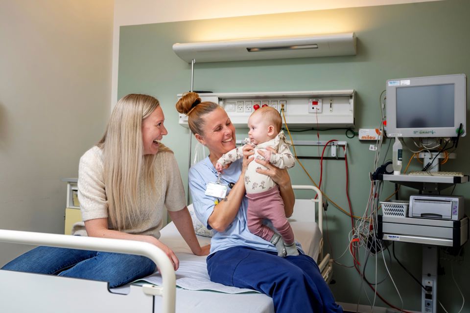 To kvinder sidder på fødestue den ene med en baby i armene. De kigger begge på babyen og smiler stort.