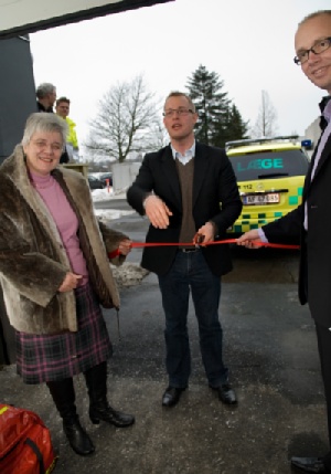 Regionsrådsformand Carl Holst (V) klipper snoren  over med et værktøj fra lægebilens taske.  En form for kniv, som bl.a. kan bruges til at klippe  sikkerhedsseler over med. Her ses han sammen  med borgmester i Kolding Kommune, Jørn Pedersen,  samt praktiserende læge Jytte Frøkjær. Det er  hende, der tidligere har kørt lægebilen fra sin  praksis i Lunderskov. 
