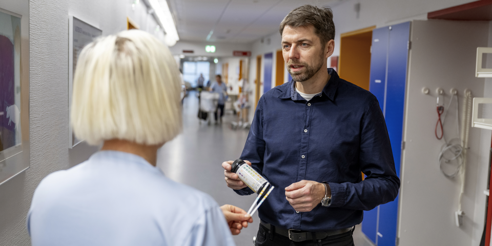 Sygeplejerske set fra ryggen i samtale med mandlig læge, som holder et glas med urinstix i hånden.